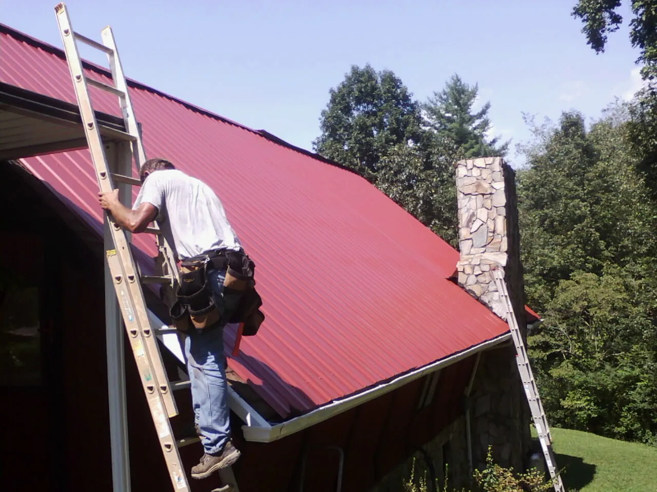 Roofer on a ladder beside a red metal roof and stone chimney — Soffit & Fascia Repair by Roofing Brothers in Ainsworth